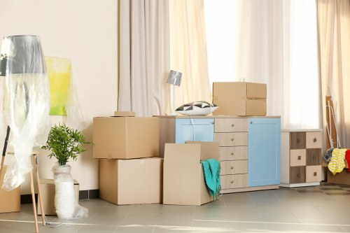 The image shows a domestic interior, likely a living room or hallway, with various household items and moving boxes arranged near a window with white curtains. In the foreground, there are several cardboard moving boxes of different sizes, some stacked and others placed on the floor, with a teal cloth draped over one box. Behind the boxes, a light blue and cream-colored wooden cabinet or sideboard is visible, with a small silver desk lamp and a vintage-style telephone on top. To the left, a tall, slim, clear glass vase with protective wrapping and a potted green houseplant in a white textured ceramic pot are positioned near the corner. The floor appears to be tiled in grey, and natural light filters through the curtains, creating a bright, neutral atmosphere. The overall scene suggests the process of packing or unpacking during a house move, with the furniture and items arranged in a clean, organized manner, consistent with house removal services provided by West Hampstead Man with Van, in the London NW6 area near Hampstead and West Hampstead.
