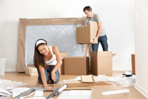 A young woman is kneeling on a wooden floor, unpacking or preparing packing materials with a roll of bubble wrap and a knife, while a young man stands nearby holding a cardboard box, both engaged in a house removal process in a bright, modern room. The background features a large, partially assembled or covered window or door frame with protective plastic sheeting, and several cardboard boxes are stacked or arranged around them. The scene illustrates a residential interior in West Hampstead during a move, with clear details of cardboard boxes, packing supplies, and casual clothing, capturing a typical house relocation environment. The lighting is natural, emphasizing the clean, uncluttered space suitable for house removals and relocation services provided by West Hampstead Man with Van.