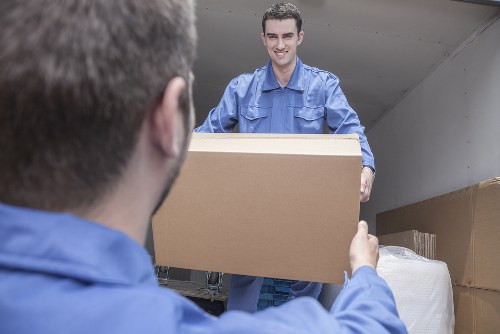 In the image, a professional house removals worker from West Hampstead Man with Van is inside a residential property, standing at the entrance of a moving van. He is wearing a blue work shirt and is smiling as he hands over a large, unmarked cardboard box to a colleague whose back is facing the camera. The interior of the van, visible in the background, contains neatly stacked moving boxes and wrapped furniture items, some covered with protective blankets. The scene takes place on a residential street, likely in or near West Hampstead, with natural daylight illuminating the space. The boxes vary in size and are made of brown cardboard, some taped at the edges, and are arranged in an organized manner. The overall setting reflects a professional and careful approach to house relocations, with attention to secure handling of household belongings during a move within London or surrounding areas.