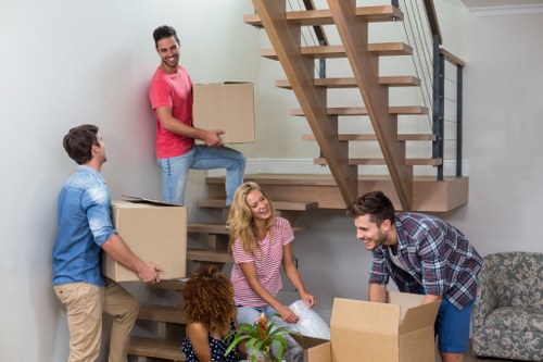 A group of five people, including three men and two women, are in a spacious living room engaged in packing and organizing items for a house move, with some holding cardboard boxes. One man in a pink t-shirt and jeans is kneeling on the staircase, holding a large box open, while another man in a blue shirt and beige trousers carries a box towards the group. A woman in a pink striped top is seated on the floor, smiling and assisting with packing, alongside a woman with curly hair and a patterned top who is also handling boxes. The living room features a modern wooden staircase with black metal railings, visible behind the group, and a comfortable grey patterned sofa on the right. The space has neutral-colored walls and ample natural light, creating a welcoming environment typical of a residential property in West Hampstead. The scene captures the activity of a house removal with careful handling of household belongings, emphasizing the professional services of West Hampstead Man with Van in household moving and removals across the local area, including the NW6 postcode district.