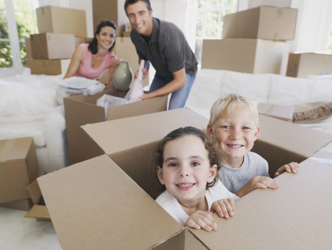 The image shows a family engaged in house removal preparations inside a bright, spacious living room with large windows allowing natural light to fill the space. In the foreground, three children are playing and smiling inside a large, open cardboard box, which is situated among other packed boxes of various sizes, some stacked on top of each other. The children have light brown and blonde hair and are dressed casually, with one girl in a white top and the other in a light grey t-shirt. Behind them, an adult man, likely a parent or removal team member, is kneeling and holding a white household lamp while smiling at the camera. To the left, a woman dressed in a pink top is kneeling beside some packed boxes, and she appears to be packing or organizing items. The room’s light-colored walls and minimal furniture create a clean, organized environment indicative of a house being prepared for relocation. The scene captures the early stages of a home move, with household items like boxes, a lamp, and other essentials ready for transportation, highlighting the professional removals services that West Hampstead Man with Van offers in the area near NW London.