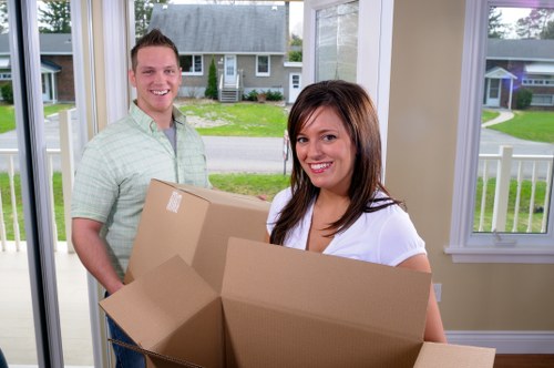 A young man and woman, both smiling, are inside a bright, well-lit conservatory or porch area of a residential property, with large windows showing a suburban street with houses and greenery outside. The woman, with shoulder-length brown hair, is holding an open cardboard moving box filled with various household items, while the man, with short light brown hair, stands behind her, also holding a cardboard box. The boxes are made of brown kraft paper and appear to contain small household goods, possibly for a house removal or move. The interior features white window frames and light-colored walls, suggesting a clean and modern home environment commonly associated with house moving services in West Hampstead or nearby areas, aligned with the services of West Hampstead Man with Van. The scene depicts the process of packing or unpacking, relevant to the company's removals service offering in the local London area, with the focus on residential relocation preparations.