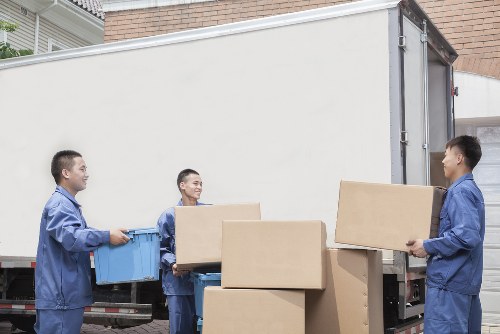 Three professional movers from West Hampstead Man with Van, dressed in blue work uniforms, are loading and unloading cardboard boxes onto and from a large white removals van parked on a residential street in Hampstead. One mover is carrying a large box on the right side, another is passing a smaller box towards the first, while the third is holding a blue plastic container. The boxes vary in size, with some stacked on the ground and others being handed between the movers. The scene takes place in front of a brick house, with a driveway and residential buildings visible in the background. The lighting suggests it is daytime with natural light illuminating the area, highlighting the textures of the cardboard and the uniforms. The image captures the careful handling and organization typical of local house removals in the Hampstead area, reflecting the services offered by West Hampstead Man with Van for efficient and professional moving solutions within London.