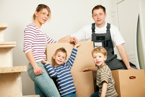 A family of four preparing for a house move, with two young children and two adults standing inside a well-lit residential space near a staircase. The mother, wearing a red and white striped long-sleeve shirt and jeans, is smiling and holding a cardboard box, while the father, dressed in a white T-shirt and black overalls, stands beside her, holding the box with one hand. The two young children, a boy and a girl, are standing in front of the boxes, both wearing striped tops; the boy is smiling and reaching upward towards the mother, and the girl looks towards the camera with a neutral expression. The scene shows several cardboard boxes stacked on the floor, and the background includes a white wall and a staircase with beige carpeted steps, suggesting an interior space of a typical UK home. The environment appears clean, organized, with natural lighting highlighting the family's activity, emphasizing the process of packing or unpacking during a house removal using professional services such as West Hampstead Man with Van, based near NW London postcode areas, focused on local house removals.
