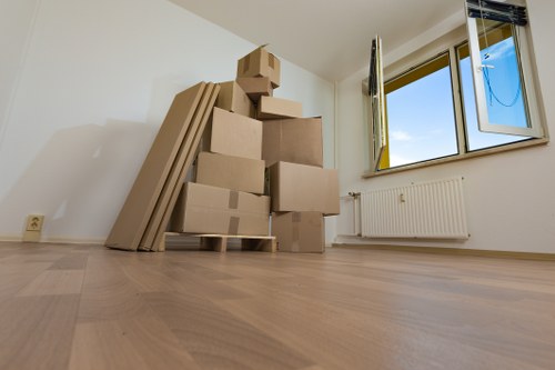 In a well-lit room with light wooden flooring and white walls, several cardboard boxes of various sizes are stacked against the left side, some arranged on a wooden pallet. To the left of the boxes, there are flat cardboard sheets leaning upright. A double-glazed window on the right wall is open, revealing a bright blue sky with some clouds, and a white radiator is positioned beneath the window. The room appears to be part of a residential property in West Hampstead, with a clean, minimal appearance suitable for a house removal or relocation process. The setting suggests an empty or newly vacated space prepared for furniture and personal belongings to be moved in or out, aligning with professional removals services by West Hampstead Man with Van aimed at sustainable and efficient house moves in the area.