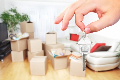 A person's hand is holding a key with a house-shaped keyring, positioned in the foreground of a living room scene. In the background, there are several cardboard moving boxes of varying sizes, some stacked on top of each other, which are typical of house removals in West Hampstead. The boxes are made of plain brown corrugated cardboard, with some showing tape closures, and contain household items, though the specific contents are not visible. The interior space features a light-colored wooden floor and a white wall with sheer curtains allowing natural light to illuminate the room. To the left, a potted green plant is partially visible, adding a natural element to the setting. In the background, a white sofa with a red throw pillow and a beige armchair are visible, complementing the cozy, lived-in atmosphere. The image subtly reflects the process of moving house, aligning with local house removal services, and captures the moment before a new chapter begins in a residential setting in West Hampstead.