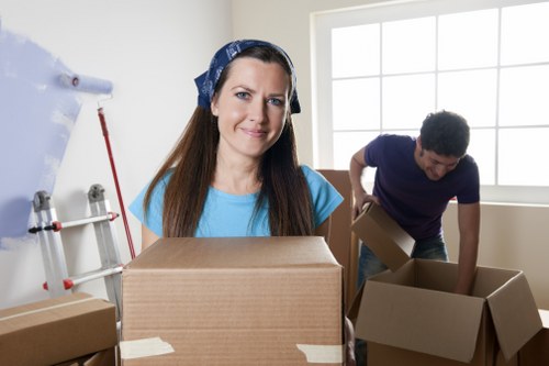 In a well-lit residential room with a large window allowing natural daylight, a woman with long brown hair, wearing a blue headscarf and matching blue top, is smiling and holding a medium-sized cardboard box with packing tape secured around it. Behind her, a man with short curly dark hair, dressed in a dark blue t-shirt, is actively engaged in packing additional boxes, one of which he is opening or filling. The room contains several cardboard boxes of varying sizes, some stacked, others placed on the floor, indicating a packing or moving process. To the left, a partial view of a wall-mounted pipe and a red-handled tool suggest the presence of household fixtures or maintenance items. The setting appears to be a home interior during a house removal, with West Hampstead Man with Van providing professional packing and moving services, subtly referencing the local area within NW postcode regions. The scene captures the essential elements of house removals, including furniture, personal belongings, and packing materials, with a focus on a professional and organized move.