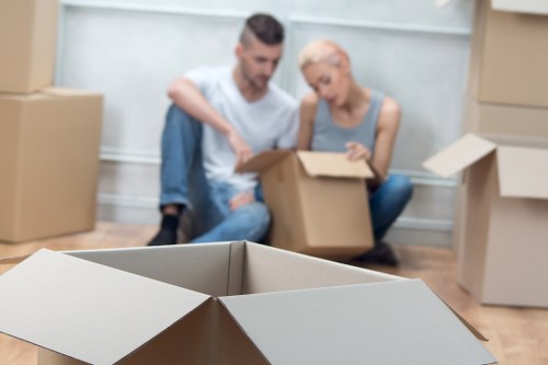 A residential interior scene showing a man and woman sitting on the wooden floor in a room with light-colored walls, surrounded by open and stacked cardboard boxes used for house removals. The man, with short dark hair and a beard, is wearing a white t-shirt and blue jeans, holding a smaller box while pointing at it. The woman, with platinum blonde hair styled in a short cut, is dressed in a sleeveless grey top, attentively looking at the box. In the foreground, an empty open cardboard box is visible, along with several other packed boxes stacked against the walls, some sealed and others open, revealing household items. The setting suggests an ongoing move within a property in or near West Hampstead, with a focus on household belongings such as cardboard boxes, typical of a house removals operation in the London area. Ambient lighting highlights the clarity of the scene, emphasizing the preparation process involved in local house relocations, as performed by West Hampstead Man with Van.