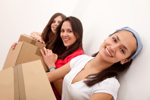 Three women with light skin and smiling expressions are standing indoors, closely together, during a house removal process. The woman in the foreground, wearing a white top and a blue hairnet, is holding a medium-sized cardboard box with both hands, positioned in front of her. Behind her, two women are partially visible; the woman in the middle, dressed in a red top, is gently holding another cardboard box, while the third woman on the left, with long brown hair, is supporting a larger cardboard box. The background features a plain white wall, and the scene appears well-lit with natural or neutral lighting. All three women are engaged in lifting or carrying boxes, indicative of a household move. The overall ambiance suggests a professional removals service environment, possibly from a property in West Hampstead or nearby London areas, aligned with the services offered by West Hampstead Man with Van specializing in removals, packing, and relocation tasks.