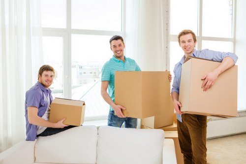 Three men are participating in a house removal in a well-lit living room, with large windows allowing natural daylight. The man on the left, dressed in a purple shirt, is holding a medium-sized cardboard box with both hands, standing next to a white sofa. The man in the middle, wearing a turquoise-striped polo shirt, is smiling and holding a large cardboard box, positioned near the center of the room. The man on the right, dressed in a blue shirt and brown trousers, is carrying a sizeable cardboard box, cradling it with one arm, and standing near a white radiator below the windows. The background reveals a bright, modern interior with a light wall and white window frames, creating a spacious and clean environment. The scene depicts a typical domestic move, with household items and boxes prepared for loading, reflecting professional house removal services in West Hampstead, London, within the N6 postcode area. All items and furniture are made of cardboard, with a focus on practical, everyday household belongings associated with residential relocation.