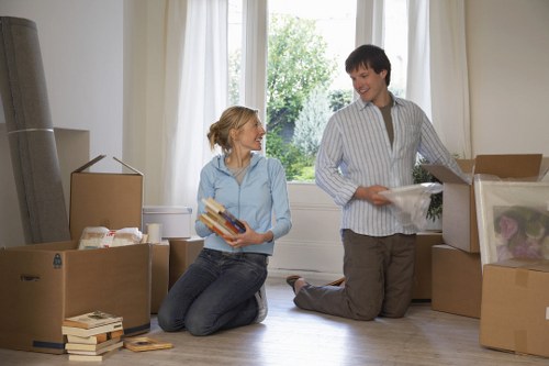 A woman and a man inside a bright residential room surrounded by moving boxes, with the woman sitting on the floor holding a stack of books and the man kneeling next to an open cardboard box, unpacking or packing items. The room features a large window with white curtains, allowing natural light to illuminate the space, and a view of green trees outside. The boxes vary in size and are filled with household items, including books, papers, and miscellaneous belongings. The woman has light-colored hair tied back and is dressed casually in a light blue shirt and dark jeans, while the man has short dark hair and is wearing a striped shirt and beige trousers. The setting appears to be part of a house or flat in West Hampstead, and the scene is indicative of a house move or relocation process managed by West Hampstead Man with Van, demonstrating careful packing and unpacking in preparation for a move within the area.