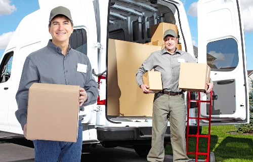 Two male removal workers from West Hampstead Man with Van are engaged in a move, with one standing outside a white removals van on a residential driveway, smiling and holding a medium-sized cardboard box. The other worker is positioned to the right, slightly behind, and is carrying a similar-sized cardboard box, while the open van's interior reveals a stacking of larger boxes, some sealed with packing tape, on a background of a suburban street with greenery and a clear sky. The men are dressed in grey uniforms with matching caps, appropriate for house removals services in the Hampstead area, and the scene depicts a professional and organized moving operation. The van's back doors are wide open, exposing the neatly packed interior with various household items prepared for transport, including furniture partially visible, all set against a neat, well-maintained residential property setting.