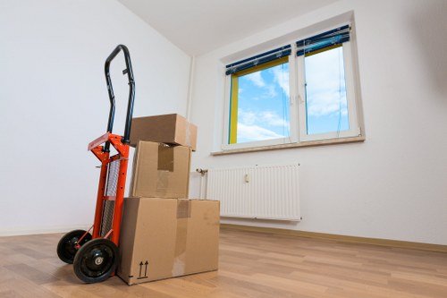 The image shows an interior scene of a sparsely furnished room with white walls and a light wooden floor. In the foreground, there are three cardboard boxes stacked on top of each other, with the smallest on top, all sealed with brown packing tape. To the left of the boxes, a red hand truck or dolly is standing upright, equipped with black wheels and a black handle. In the background, a white radiator is mounted on the wall beneath a double-glazed window that has a white frame and blue blinds partially raised, revealing a blue sky with some white clouds outside. The room appears well-lit with natural daylight, suggesting a residential setting that could be part of a house or apartment in West Hampstead, London, a typical area served by house removals companies such as West Hampstead Man with Van. The scene conveys a moment of packing or moving preparations, emphasizing the readiness for a house relocation or professional removal service within the local area.
