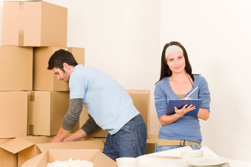 A man and a woman are in a residential setting, likely a house in West Hampstead, preparing for a move as part of a removals service. The man, wearing a light blue T-shirt and dark jeans, is focused on packing and organizing a collection of cardboard boxes, some of which are stacked against a white wall. He is leaning forward, handling an open box with packing material visible inside. The woman, dressed in a grey long-sleeve shirt and beige trousers, is standing nearby holding a clipboard or tablet, appearing to coordinate or check items. She has a neutral expression and is wearing a headband. In the foreground, on a white surface, there are several white bowls and plates, indicating this could be a kitchen or dining area. The environment is well-lit with neutral tones, emphasizing a professional and organized scene related to house moving services. The overall context suggests a carefully managed domestic relocation, typical of West Hampstead's residential neighbourhoods.