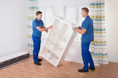 Two professional movers dressed in blue uniforms are inside a bright, minimally decorated room with light wooden flooring and white walls. They are carefully handling a white wooden shelving unit, lifting and positioning it between them. The shelving unit features multiple open compartments, and its surface appears smooth with a polished finish. Behind the movers, there is a large window with white blinds that are partially open, allowing natural light to illuminate the space. To the right, a floor-length curtain with horizontal stripes in shades of green, blue, and beige hangs on a rod. On the left side of the room, a slim black radiator is installed along the wall. The overall setting suggests a residential interior, possibly in a house or apartment in West Hampstead, and the scene depicts part of a domestic move or furniture installation, with [West Hampstead Man with Van] providing professional removals services in the area.