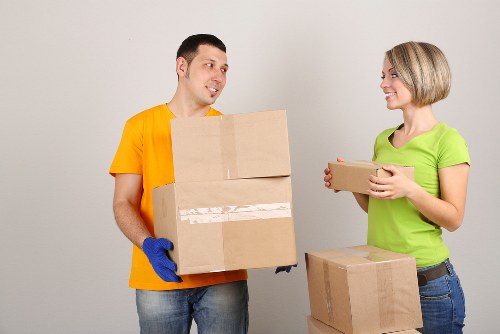 A professional house removals team member from West Hampstead Man with Van is seen indoors, wearing a bright yellow T-shirt and blue gloves, carrying a medium-sized cardboard box with clear packing tape along the edges. He appears to be engaged in conversation with a woman, who is smiling and wearing a light green T-shirt and blue jeans, holding a smaller, flat-packed cardboard box. On the floor beside her, there is an additional larger box, also made of cardboard and sealed with packing tape. The setting is a neutral, well-lit interior space, likely part of a residential property, with a plain light grey wall in the background. The scene depicts the process of packing or unpacking, with household items and boxes arranged naturally for a house move. The focus is on the careful handling and organization of belongings during a local relocation service, representative of West Hampstead's household removals activities, emphasizing safety and professionalism during the process.