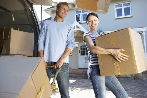 A man and a woman, both smiling, are engaged in a house removal outside a residential property. The man, dressed in a light blue long-sleeved shirt and jeans, is holding a dolly with handles, assisting in transporting large cardboard boxes. The woman, wearing a striped t-shirt and jeans, is carrying a sizable cardboard box with visible brown tape securing it, positioned in front of her as she moves toward the open rear of a large grey removals van parked on a driveway. The van’s interior shows additional cardboard boxes, indicating a loading process for a house move within West Hampstead or nearby London. The background reveals a residential street scene with a two-storey house featuring white-framed windows and a front porch. The scene is well-lit by natural daylight, emphasizing the casual, professional atmosphere of a house removal team. The image captures the practical and organized nature of a local removals service associated with West Hampstead Man with Van, highlighting their role in residential relocations in the area.
