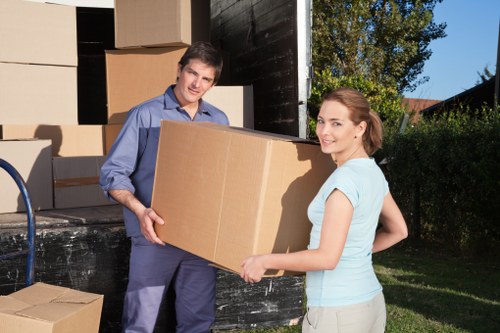 Two professional movers from West Hampstead Man with Van are outside a residential property, loading large cardboard boxes into a black removals van parked on a driveway. The man, wearing a blue shirt and dark trousers, is holding the box from one side while the woman, dressed in a light blue top and beige trousers, supports it from the opposite side, smiling at the camera. The background features a garden with trees and bushes, as well as the house's exterior wall and window. Several other packed boxes of various sizes, some wrapped in brown packing paper, are stacked near the van and on the ground, indicating a house removal in progress. The scene is set in bright daylight, with natural shadows suggesting clear weather, highlighting the careful handling of household items for a safe move within West Hampstead or nearby areas in London.