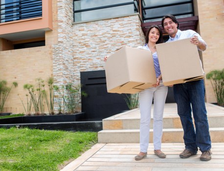 A young couple standing outside a modern residential property in West Hampstead, each holding a large cardboard moving box. The woman has shoulder-length dark hair, wearing a light blue shirt and white trousers, smiling at the camera. The man has short dark hair, wearing a white shirt and dark jeans, also smiling. The background features a contemporary house with a mix of brick and stone exterior, a small garden with green grass, potted plants, and a pathway leading to the entrance. The scene appears to be part of a house move, with the couple prepared for relocation, and is indicative of professional removals services in West Hampstead, London, with the focus on household items such as boxes and the residential setting.