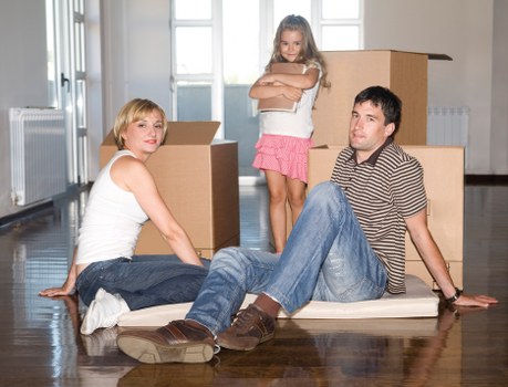 The image depicts a family in a well-lit room with wooden flooring, surrounded by several large cardboard moving boxes, indicating a house relocation. Two adults, a woman with short blonde hair wearing a white sleeveless top and blue jeans, and a man with dark hair dressed in a striped polo shirt and blue jeans, are seated on the floor, leaning slightly back with their legs extended and hands supporting them. Between them, a young girl with long, light brown hair, dressed in a pink skirt and white top, stands with arms crossed, observing the scene. The background shows a white radiator, a window with closed blinds, and a plain wall, adding to the residential atmosphere. The scene suggests completion of packing or preparation for a move, consistent with services offered by West Hampstead Man with Van in the Westminster area, reflecting a typical house removal scenario within London or nearby communities. Natural lighting enhances the clarity of household items and their textures, such as the cardboard boxes and the family's casual attire, conveying a moment of pause during a household moving process.