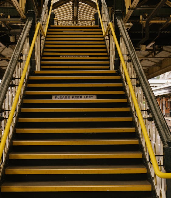 A set of metal stairs with wooden treads featuring yellow safety edges, located inside a transport or warehouse facility. The stairs are flanked by yellow handrails on both sides and lead up to an ele